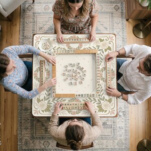 May include: A square mahjong table featuring a cream-colored surface with a decorative border of green leaves and pink roses. The table is set with a central pile of mahjong tiles, four tile racks, and four players seated around the perimeter.