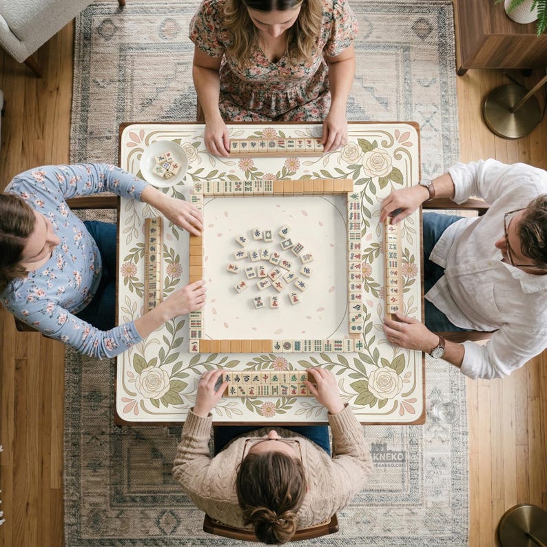 May include: A square mahjong table featuring a cream-colored surface with a decorative border of green leaves and pink roses. The table is set with a central pile of mahjong tiles, four tile racks, and four players seated around the perimeter.