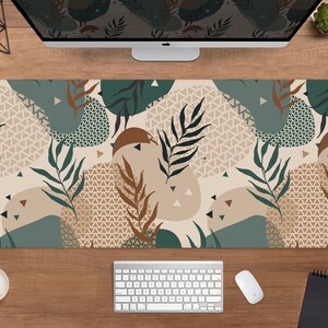 May include: A rectangular desk mat featuring a botanical design with green, brown, and beige abstract shapes and leaf patterns. The mat is shown on a wooden desk with a computer monitor, keyboard, and mouse.