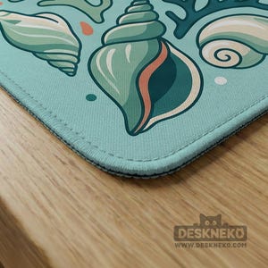 May include: A light blue desk mat with a seashell design in teal, green, and coral. The mat has a stitched edge and a repeating pattern of various seashells and coral. The Deskneko logo is visible in the bottom right corner.