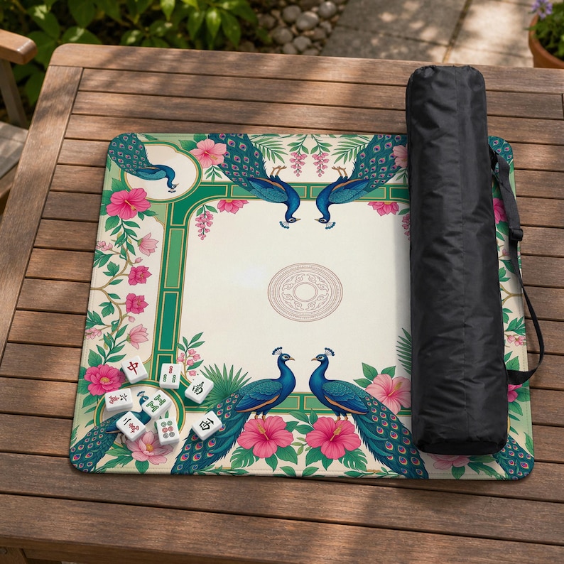 May include: A square mahjong mat featuring a cream-colored center with a circular emblem, surrounded by a green border decorated with blue peacocks and pink hibiscus flowers. A small pile of white mahjong tiles and a black carrying case are included.