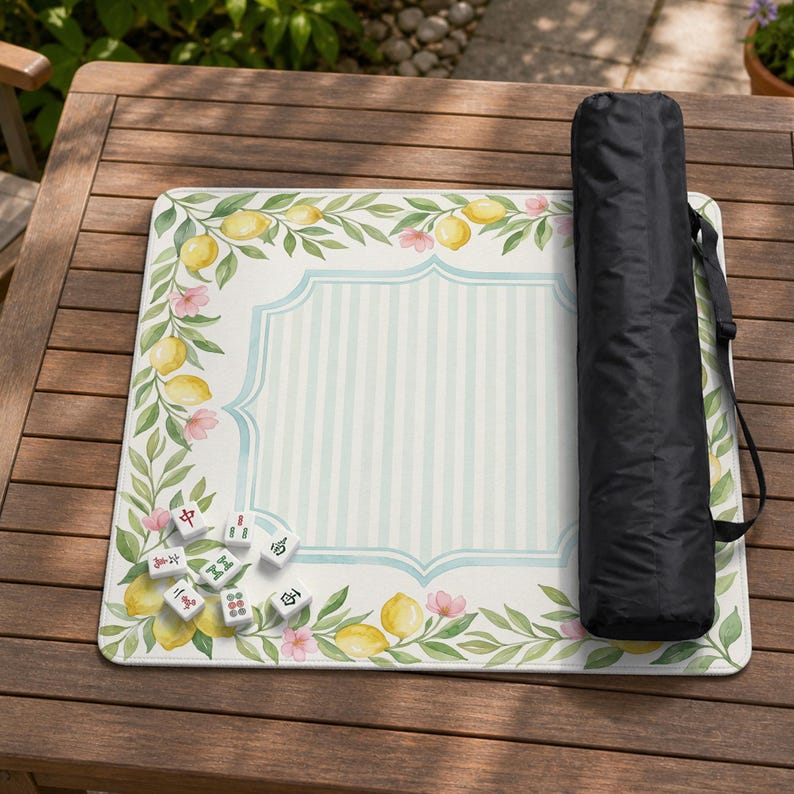 May include: A rectangular mahjong mat with a lemon and floral design, a light blue striped center, and a black carrying case. Several mahjong tiles are scattered on the mat. The mat is on a wooden table.