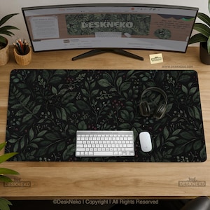 Puede incluir: Una alfombrilla de escritorio negra con un patrón botánico de hojas verdes y bayas rojas. Un teclado y un ratón blancos están sobre la alfombrilla, junto con unos auriculares. El monitor muestra el sitio web DeskNeko.