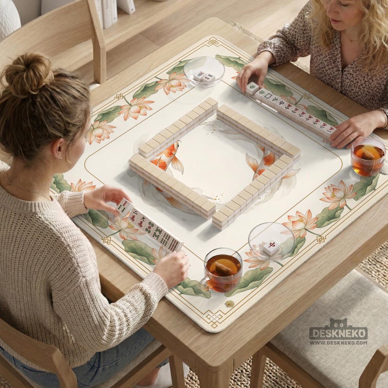 May include: A square wooden table with a cream-colored Mahjong game mat featuring a floral and koi fish design. Two people are playing Mahjong, with tiles and tea cups visible. The mat has a decorative border and the text "DESKNEKO" is visible.
