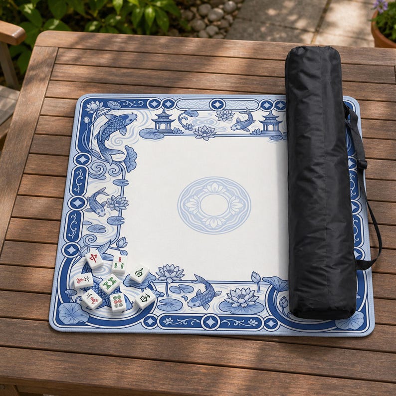 May include: A square mahjong mat featuring a blue and white pattern of koi fish, lotus flowers, and pagodas, with a central circular motif. Seven white mahjong tiles are placed on the mat, accompanied by a black cylindrical carrying case.