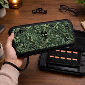 May include: Black carrying case with a green leaf pattern and black cat illustrations. The case is open, revealing a black game console and storage for game cartridges. The brand name DESKNEKO is printed on the case.