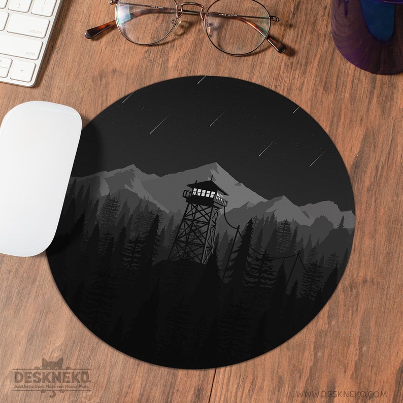 May include: Round black mouse pad featuring a grayscale illustration of a forest fire lookout tower, mountains, and a starry night sky. The mouse pad has the text "DESKNEKO" and "www.deskneko.com".