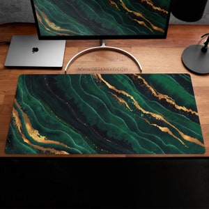May include: A large rectangular desk mat with a dark green and gold abstract design. The pattern resembles flowing waves with gold accents. The website address deskneko.com is visible in the top left corner.