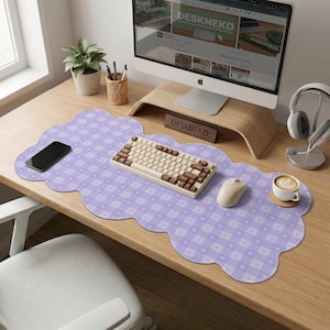 May include: A desk setup featuring a light purple cloud-shaped desk mat with a checkered pattern. A cream-colored keyboard with brown keys sits on the mat, along with a mouse, phone, and a cup of coffee. The monitor displays the text "DESKNEKO".