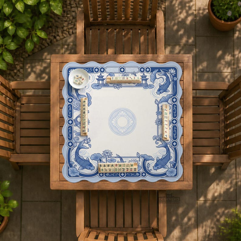 May include: A square mahjong mat with a scalloped edge featuring a blue and white pattern of koi fish, lotus flowers, and pagodas. The mat includes four wooden tile racks and a small white bowl containing mahjong tiles on a wooden table.