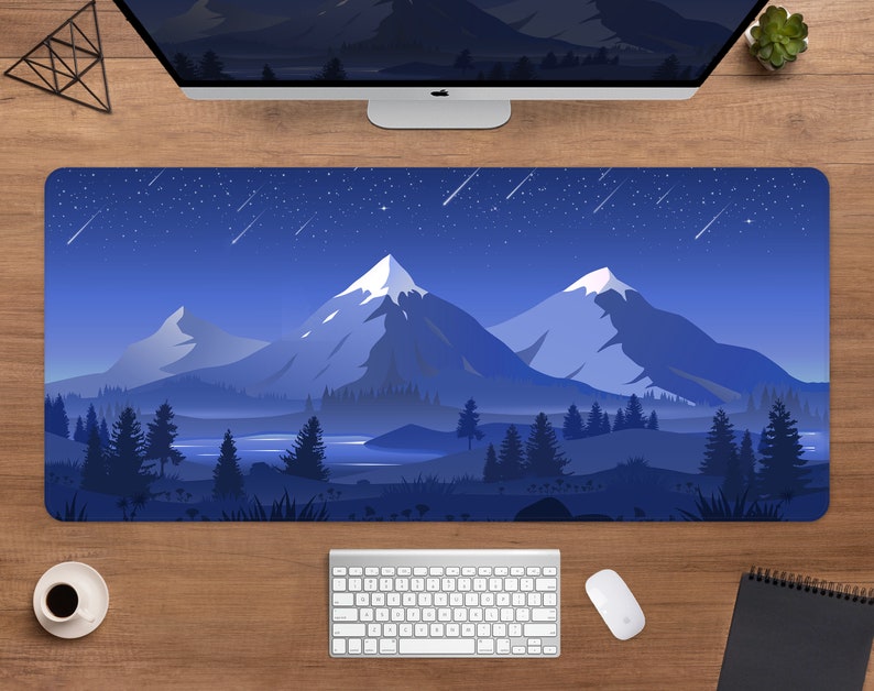 May include: A rectangular desk mat featuring a mountain landscape under a starry night sky. The design includes snow-capped peaks, a forest, and a lake, all in shades of blue. A keyboard, mouse, and coffee cup are visible.