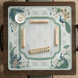 Könnte beinhalten: Ein quadratischer Mahjong-Spieltisch mit hellblauer Spielfläche mit Pfau-Illustrationen und floralen Akzenten. Der Tisch enthält Holzfliesen, Würfel und eine kleine weiße Schüssel. Der Tisch steht auf einem dunkelbraunen Holzrahmen.