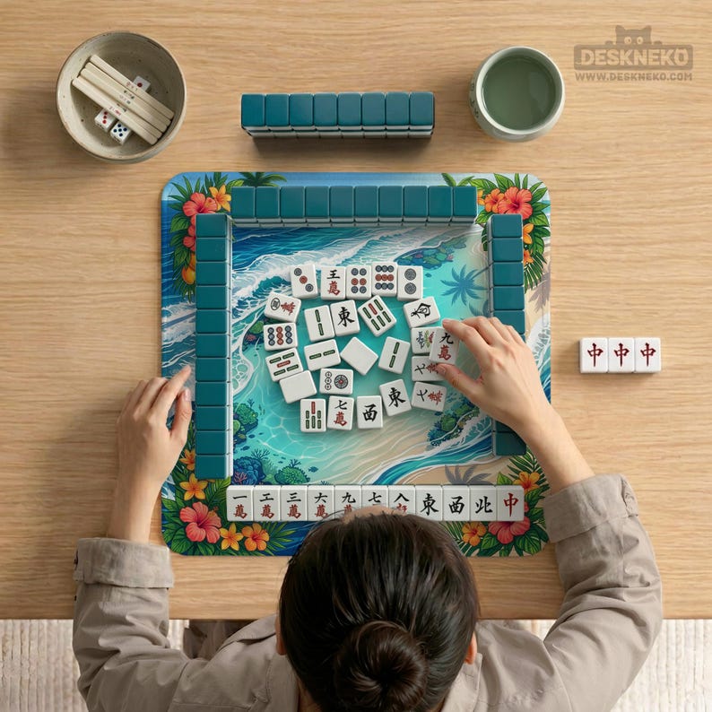 May include: A person playing Mahjong on a wooden table. The game set includes white tiles with Chinese characters, teal tile racks, dice, and a small bowl. The game board features a beach scene with floral accents.
