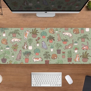 Puede incluir: Alfombrilla de escritorio rectangular con un patr&oacute;n repetido de gatos de dibujos animados y plantas en macetas sobre un fondo verde salvia. Los gatos est&aacute;n en varias poses, y las plantas est&aacute;n en macetas de terracota. Se ve una computadora, un teclado y un rat&oacute;n.