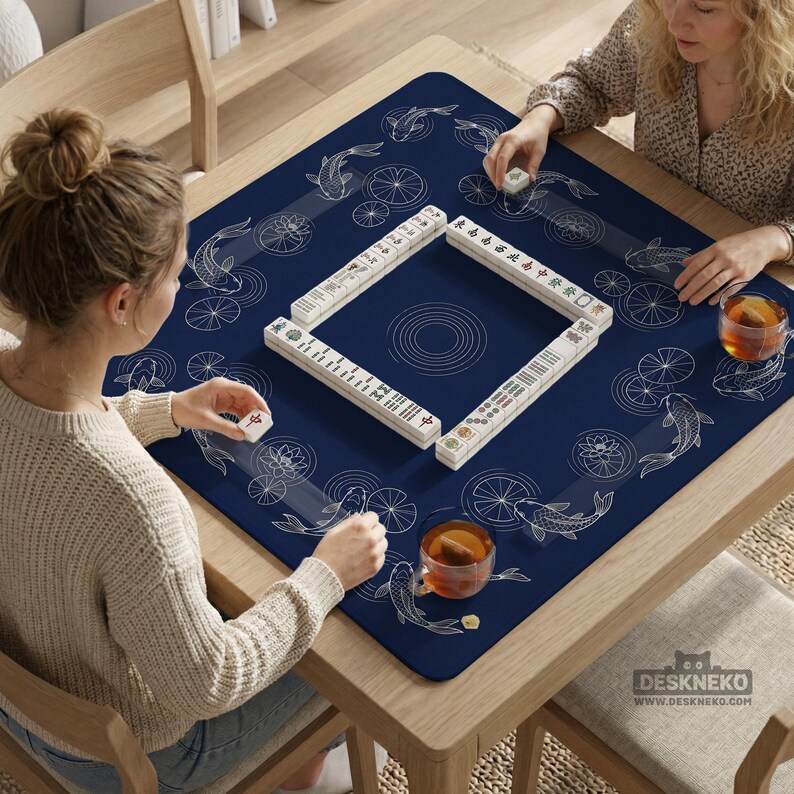 Puede incluir: Una mesa cuadrada de madera con un tapete azul marino con un dise&ntilde;o de peces koi y nen&uacute;fares. Las fichas blancas de Mahjong est&aacute;n dispuestas en un cuadrado, con dos personas jugando. Dos vasos de t&eacute; est&aacute;n sobre la mesa.