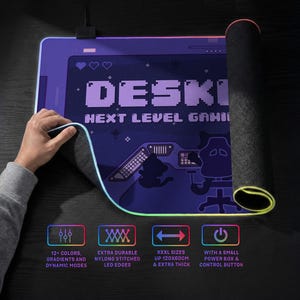 Lifestyle product image of an RGB LED desk mat on a desk, shown partially rolled to reveal the textured non-slip rubber base and 4mm thick construction. A hand lifts the corner to highlight the flexible illuminated edge with durable nylon stitching. The surface of the extra large mousepad features a high resolution print of the DESKNEKO logo. A small power box and cable are visible at the top. Icons below indicates that it's gaming grade quality and available in extra-large sizes up to 120×60 cm