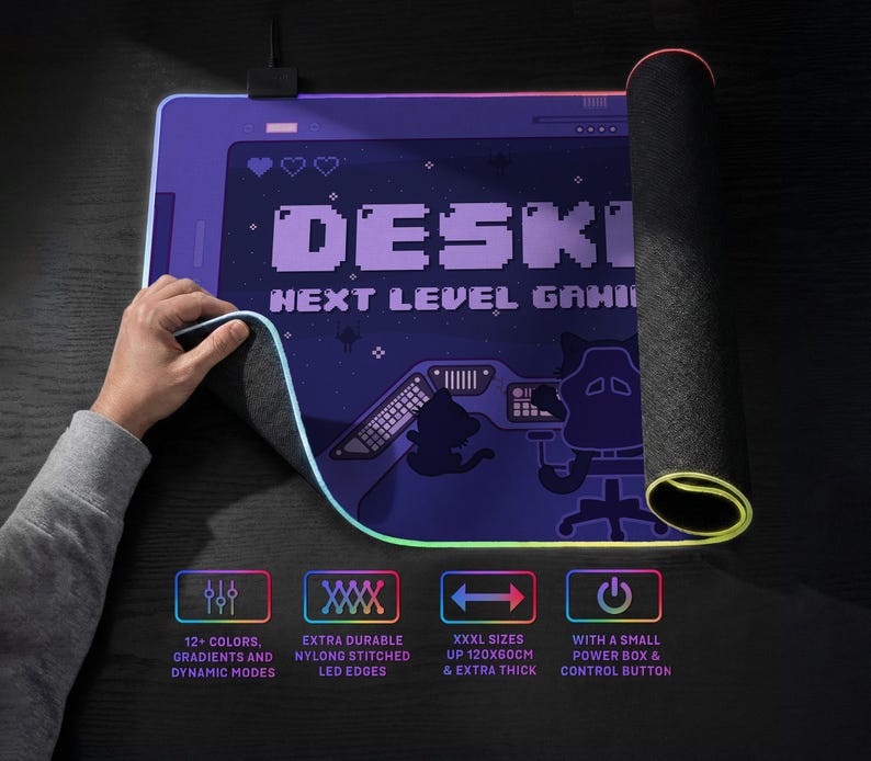 Lifestyle product image of an RGB LED desk mat on a desk, shown partially rolled to reveal the textured non-slip rubber base and 4mm thick construction. A hand lifts the corner to highlight the flexible illuminated edge with durable nylon stitching. The surface of the extra large mousepad features a high resolution print of the DESKNEKO logo. A small power box and cable are visible at the top. Icons below indicates that it's gaming grade quality and available in extra-large sizes up to 120×60 cm