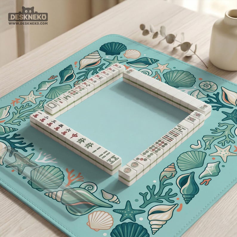 May include: A Mahjong set with white tiles and Chinese characters is arranged on a light blue mat. The mat features a sea-themed design with seashells, starfish, and coral. The game is set on a light wooden surface.