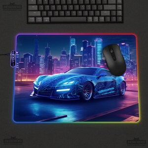 May include: A rectangular gaming mouse pad with a blue sports car design, a black mouse, and a black keyboard. The mouse pad has a colorful illuminated border and a cityscape background.
