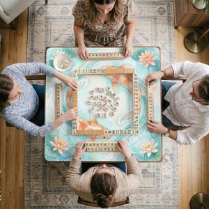K&ouml;nnte beinhalten: Ein quadratischer Mahjong-Tisch mit einer hellblauen Oberfl&auml;che, die mit orangefarbenen Koi-Fischen und rosa Lotusbl&uuml;ten illustriert ist. Der Tisch ist mit einem zentralen Stapel el elfenbeinfarbener Spielsteine und vier Ablageleisten best&uuml;ckt.