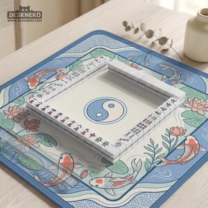 May include: A Mahjong set with white tiles and a Yin Yang symbol in the centre. The set is on a blue mat featuring koi carp and lotus flowers. The brand name "DESKNEKO" is visible. The mat's design is reminiscent of traditional Asian art.