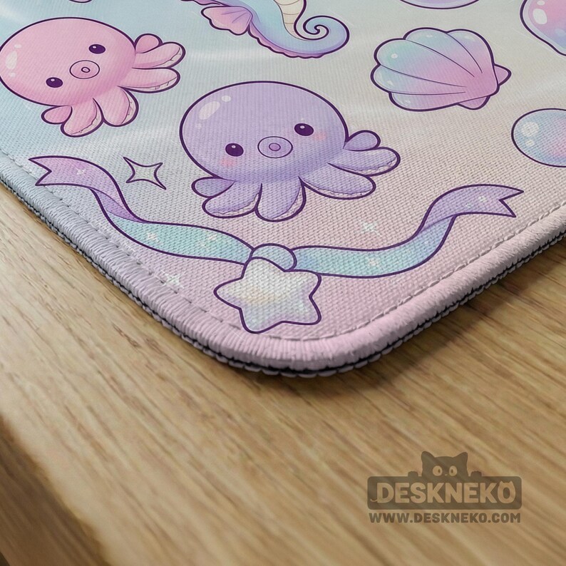 May include: A pastel-colored mousepad featuring cartoon sea creatures, including octopuses, a seahorse, and a seashell. A ribbon with a star is at the bottom. The mousepad has a black border and the Deskneko logo.