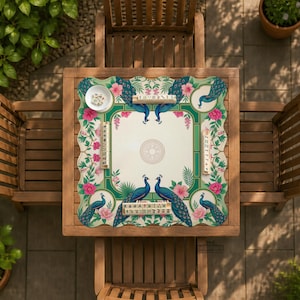 May include: A square mahjong mat featuring a cream-colored center with a circular emblem, surrounded by a green border decorated with blue peacocks, pink hibiscus flowers, and green foliage. Four sets of mahjong tiles are placed on the mat.