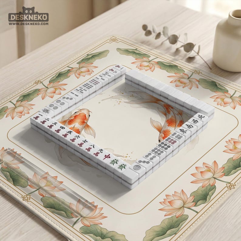 May include: A white Mahjong set arranged in a square on a cream-colored mat. The mat features a floral design with lotus flowers and koi fish. The tiles have red and green characters. The logo "DESKNEKO" is in the top left corner.