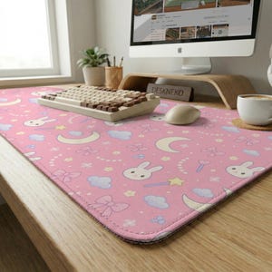 Può includere: Tappetino per scrivania rosa con un motivo ripetuto di coniglietti bianchi, lune, stelle, fiocchi e nuvole. Una tastiera e un mouse sono sul tappetino. Il tappetino ha un bordo cucito e il testo "DESKNEKO" è visibile.