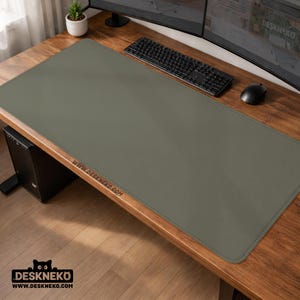 May include: An olive-green desk mat with stitched edges, on a wooden desk. A black keyboard, mouse, and computer monitor are visible. The website address "WWW.DESKNEKO.COM" is printed on the mat. The Deskneko logo is in the bottom left corner.