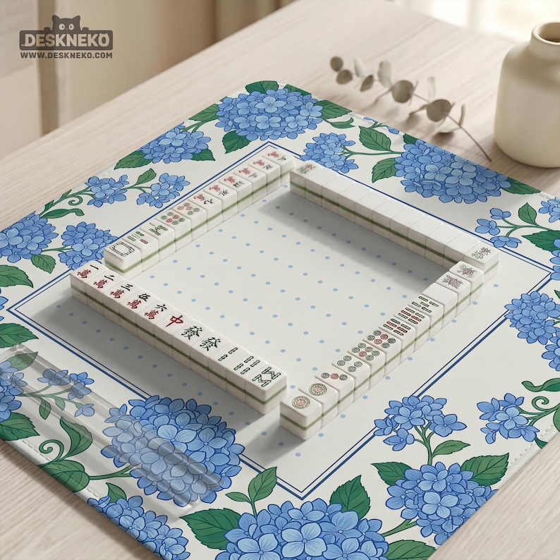 May include: A Mahjong set arranged on a floral patterned mat. The mat features blue hydrangea flowers and a white background with blue polka dots. The Mahjong tiles are white with red and green markings.