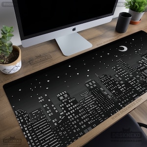 Op de afbeelding: Zwart en grijze bureauonderlegger met een gepixeld stadslandschapontwerp, een halve maan en sterren. De mat heeft de tekst "Tokyo" en andere Japanse tekst. De rechthoekige mat bedekt een houten bureau.