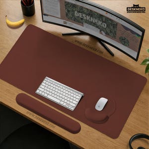 Puede incluir: Una alfombrilla de escritorio burdeos con reposamuñecas y alfombrilla de ratón a juego. Un teclado y un ratón blancos están sobre la alfombrilla. El monitor curvo muestra el texto "DESKNEKO" y la dirección del sitio web. También se ve un plátano y un portalápices.