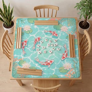 May include: A square mahjong mat featuring a light blue water surface pattern with koi fish, pink lotus flowers, and green leaves. Four wooden tile racks are placed on the edges of the mat, which is centered on a wooden table with mahjong tiles.