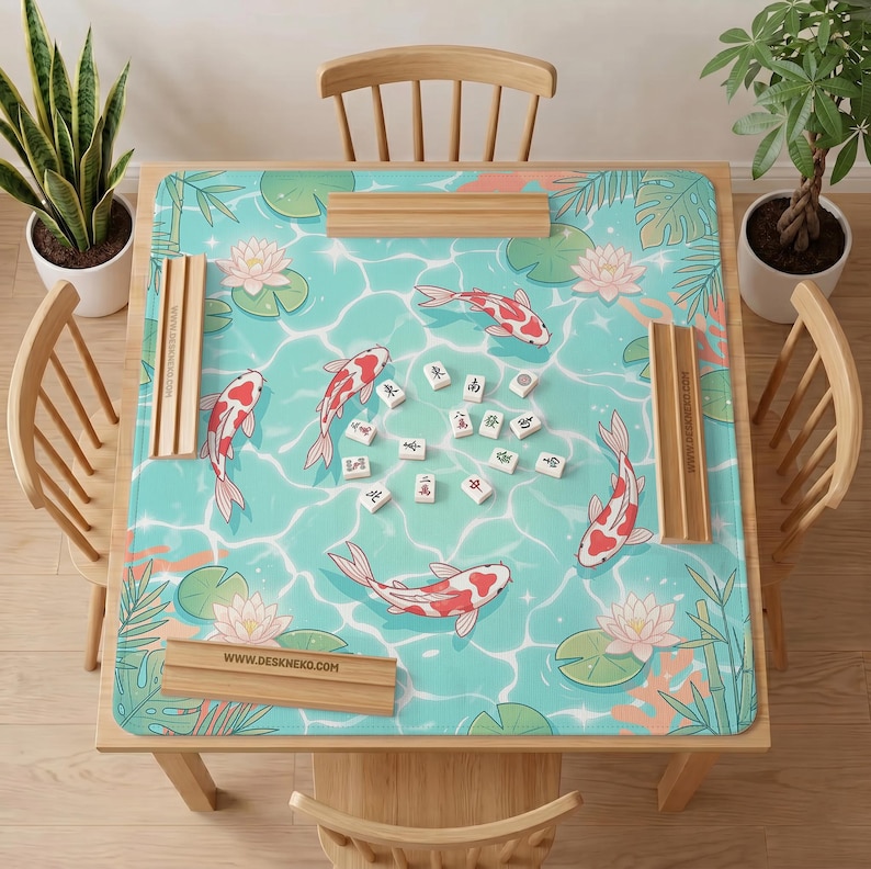 May include: A square mahjong mat featuring a light blue water surface pattern with koi fish, pink lotus flowers, and green leaves. Four wooden tile racks are placed on the edges of the mat, which is centered on a wooden table with mahjong tiles.