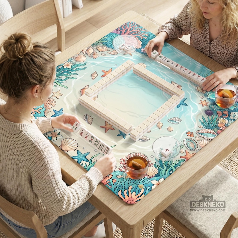 May include: A wooden mahjong set with beige tiles is set up on a table with a colorful ocean-themed mat. The mat features illustrations of seashells, starfish, and coral. Two people are playing the game, with cups of tea nearby.