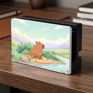 Puede incluir: Base de Nintendo Switch negra con un panel frontal decorativo. El panel presenta un capibara de dibujos animados con un pañuelo naranja, sobre un paisaje de río, colinas y cielo. La base está sobre una superficie de madera.