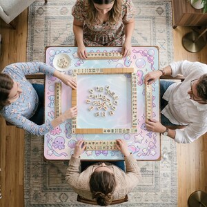 May include: Overhead view of a Mahjong game in progress. Four people are seated around a table with a pastel-colored mat featuring sea creatures and bows. Wooden tiles are arranged on the table, and each player holds a rack of tiles. The table is set on a patterned rug.
