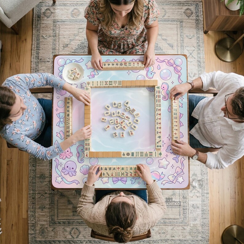 May include: Overhead view of a Mahjong game in progress. Four people are seated around a table with a pastel-colored mat featuring sea creatures and bows. Wooden tiles are arranged on the table, and each player holds a rack of tiles. The table is set on a patterned rug.