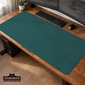 Op de afbeelding: Een teal bureaumat op een houten bureau. De mat is rechthoekig en bedekt het grootste deel van het bureaublad. Een zwart toetsenbord en muis liggen op de mat. Het websiteadres www.deskneko.com staat op de mat.