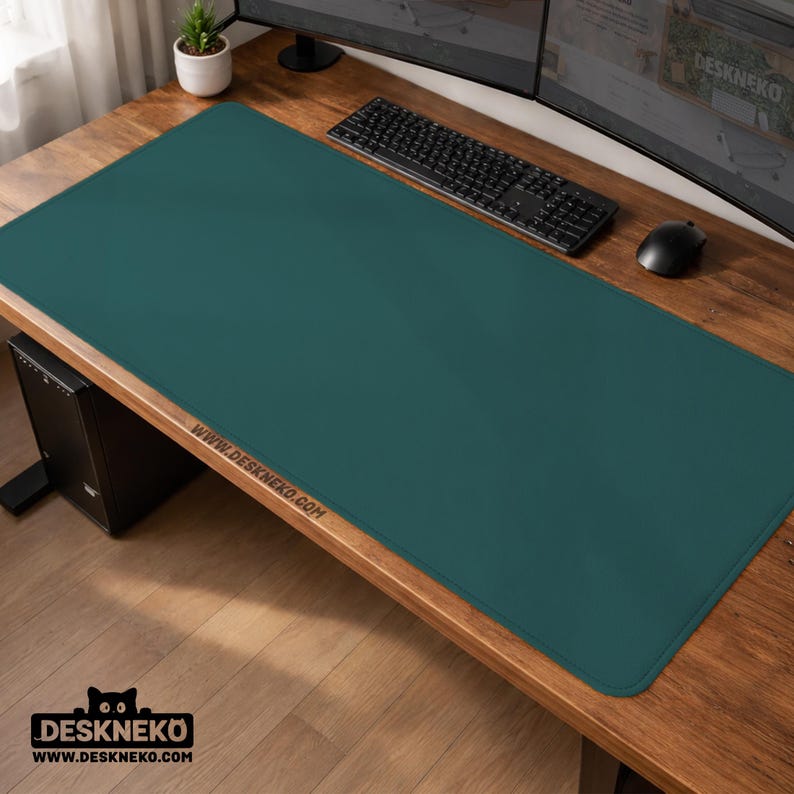 Op de afbeelding: Een teal bureaumat op een houten bureau. De mat is rechthoekig en bedekt het grootste deel van het bureaublad. Een zwart toetsenbord en muis liggen op de mat. Het websiteadres www.deskneko.com staat op de mat.