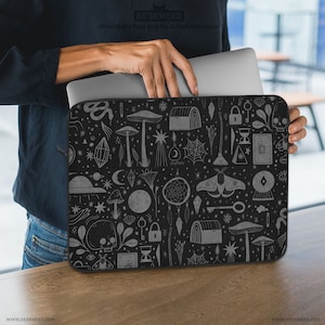 May include: Black laptop sleeve with a repeating pattern of mystical symbols, including mushrooms, crystals, keys, and a skull. The sleeve is held open, revealing a silver laptop inside. The text "DESKNEKO" is visible at the top and bottom of the sleeve.