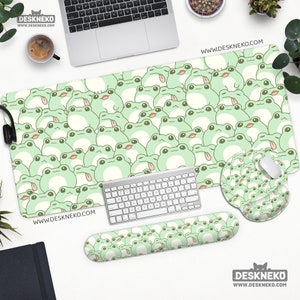 May include: A desk setup featuring a large mouse pad, mouse wrist rest, and keyboard wrist rest, all adorned with a repeating pattern of light green cartoon frogs. A matching mouse pad and a keyboard are also present. The website address www.deskneko.com is visible.