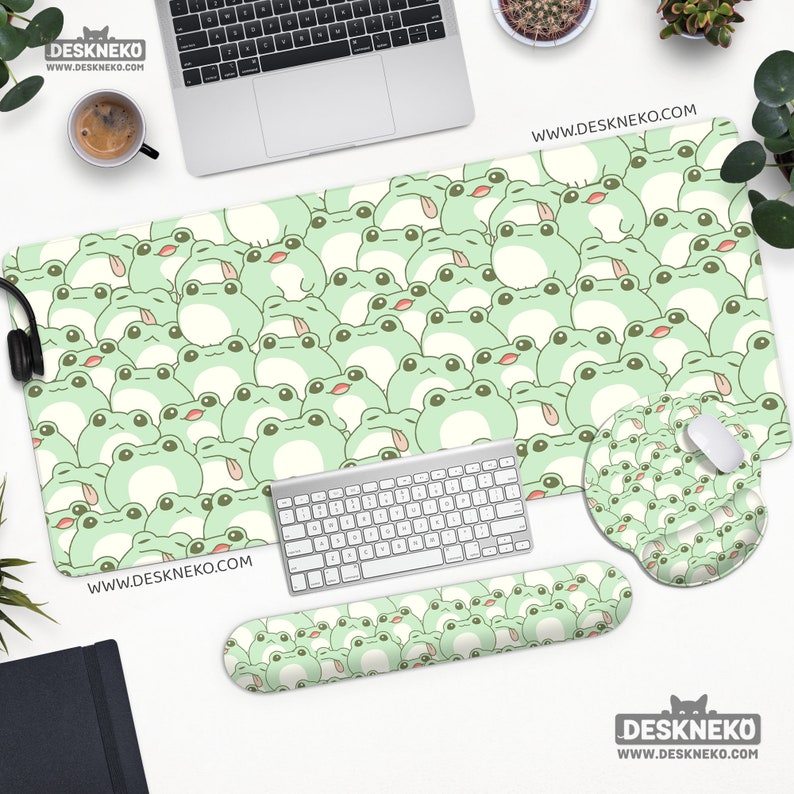 May include: A desk setup featuring a large mouse pad, mouse wrist rest, and keyboard wrist rest, all adorned with a repeating pattern of light green cartoon frogs. A matching mouse pad and a keyboard are also present. The website address www.deskneko.com is visible.