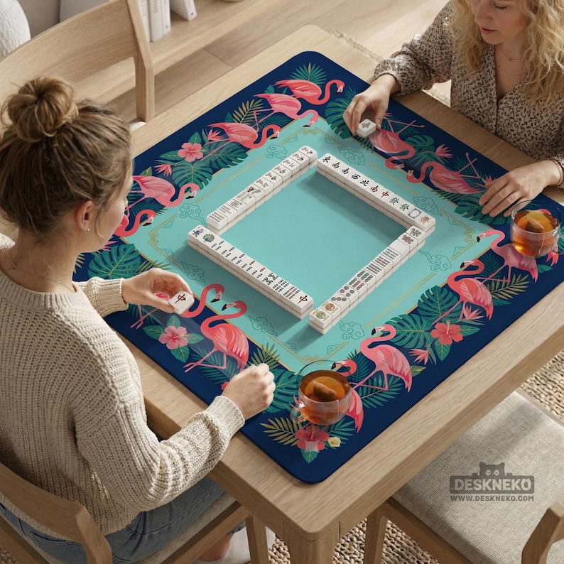 May include: A square Mahjong game mat with a flamingo and tropical leaf design. The mat is navy blue with a teal center and pink flamingos. Mahjong tiles are arranged on the mat, and two people are playing the game.