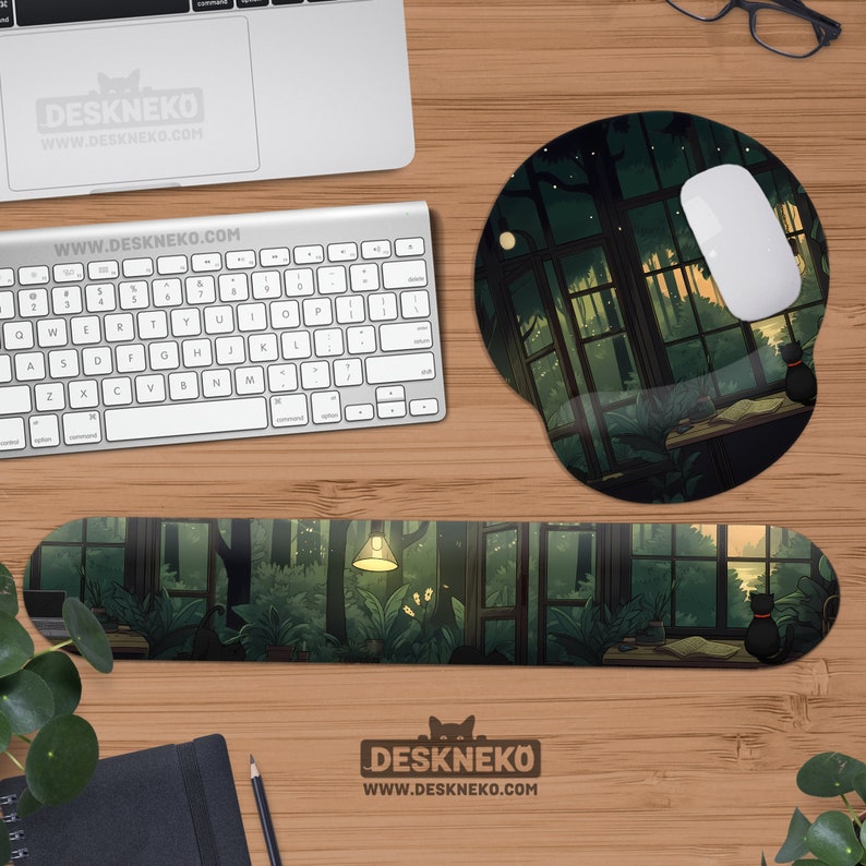 May include: A computer desk setup featuring a mousepad, wrist rest, and keyboard. The mousepad and wrist rest have a forest scene design with a black cat. The keyboard is silver. The desk setup is on a wooden surface. The brand name DESKNEKO is visible.