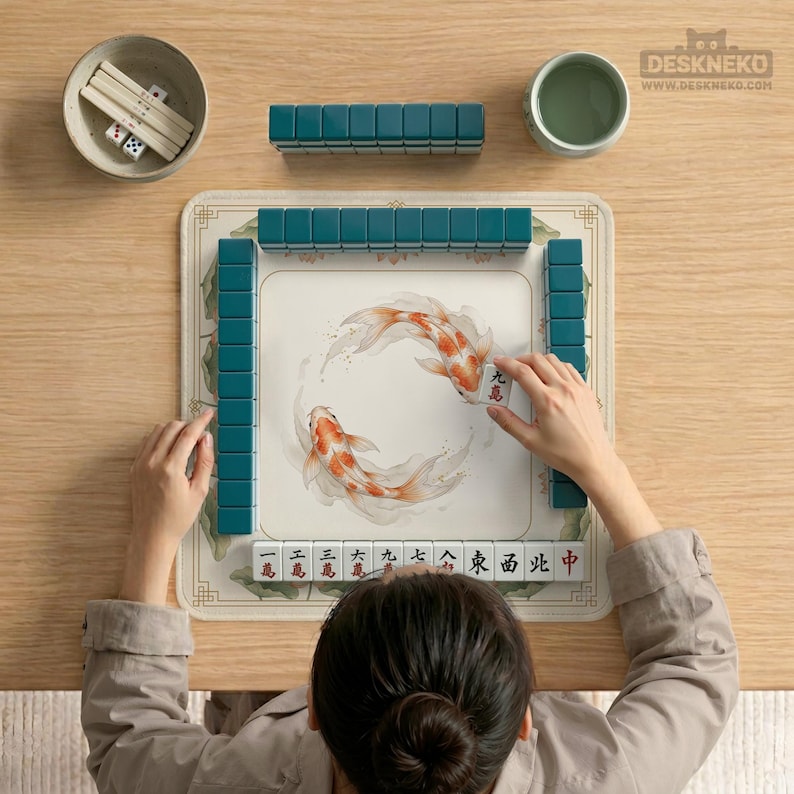 May include: A person playing Mahjong. The game set includes teal tiles, dice, and a bowl of chopsticks. The playing surface features a koi fish design. The game is set on a light wood table.