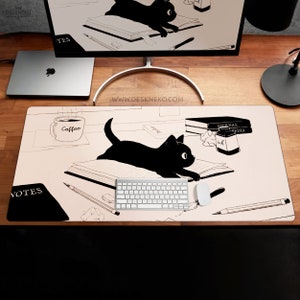 Op de afbeelding: Bureaumat met een illustratie van een zwarte kat op een open boek. Het ontwerp bevat een koffiekopje, potloden en de woorden "Journal Media" en "Notes". Een toetsenbord en een muis zijn ook zichtbaar.