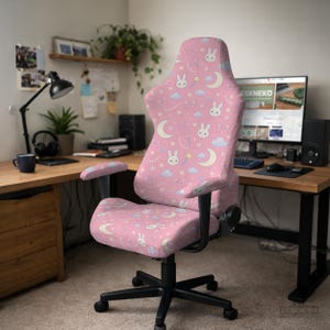 May include: A pink gaming chair featuring a repeating pattern of white rabbit faces, yellow crescent moons, light blue clouds, pink bows, and small stars. The chair has black armrests, a black five-point base with casters, and a high backrest.