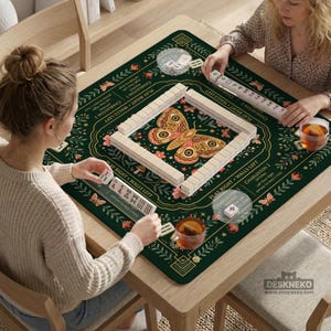 May include: A square dark green mahjong mat featuring a central moth illustration, floral patterns, and printed game instructions. The mat includes text such as Charleston 1, Charleston 2, Play Right, and Walls Left, with white tiles arranged in a square.